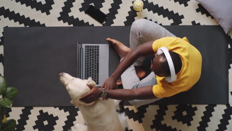Afroamerikanischer-Mann-Sitzt-Mit-Laptop-Und-Streichelhund-Auf-Dem-Boden